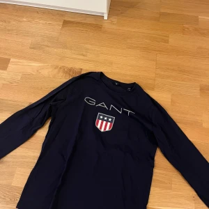 Mörkblå långärmad GANT tröja - Snygg mörkblå långärmad tröja från GANT med stort tryck av logga och sköld framtill. Tröjan är i mjuk bomull och har en klassisk passform med rund hals. Perfekt för dig som gillar stilrena och sportiga plagg med preppy känsla. Tröjan är även tunn.