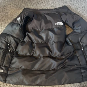 Svart dunväst från The North Face - Säljer en svart dunväst från The North Face med hög krage och två dragkedjeförsedda fickor. Västen har broderad logga på bröstet och siffran 700 på nederdelen. Perfekt för lager-på-lager och riktigt snygg till streetwear. Passar mig som är 160, men passade mig när jag var 150cm också.