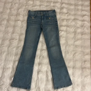 Bootcut jeans i ljusblå denim - Snygga bootcut jeans i ljusblå denim med två framfickor med lock och knappar. Klassisk femficksmodell med utsvängda ben och normal midja. Perfekta för dig som gillar retrovibbar och vill ha en avslappnad men trendig look.
