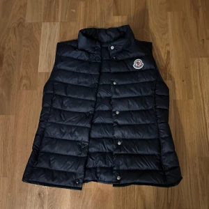 Svart dunväst från Moncler - Snygg svart dunväst från Moncler med klassisk quiltad design och hög krage. Västen har tryckknappar framtill och Monclers ikoniska logga på bröstet. Perfekt för lager-på-lager och ger en clean look till din outfit.