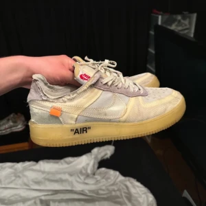 Nike Air Force 1 x Off-White sneakers - Nike Air Force 1 x Off-White sneakers i en ljus beige och vit färg med genomskinliga paneler, klassisk snörning och Off-White detaljer som orange tag och textprint på sidan. Sulan är gulaktig och har 'AIR' tryckt på sidan. Snygg och unik design för dig som gillar streetwear.