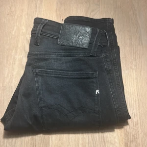 Replay Anbass svarta jeans, stl 29 - Svarta Replay Anbass jeans med klassisk femficksdesign och snygga detaljer på bakfickorna. Slimmad passform och midwaist. Jeansen är tillverkade i stretchigt bomullsmaterial för extra komfort. Perfekta för dig som gillar stilrena och moderna jeans.