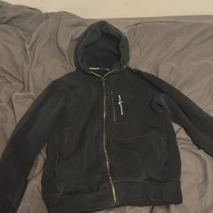 Svart hoodie från Sail Racing med dragkedja - Svart hoodie från Sail Racing i storlek M. Tröjan har hel dragkedja framtill, huva och en bröstficka med Sail Racing-logga. Perfekt för chill dagar eller när du vill ha en sportig look. Materialet känns mjukt och skönt mot huden. 