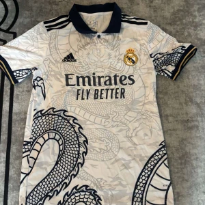 Real Madrid fotbollströja  - Snygg Real Madrid fotbollströja från Adidas i vitt med mörkblå detaljer och coolt drakmönster över hela tröjan. Klassisk krage, broderat klubbmärke och sponsortryck på bröstet. Tillverkad i lätt och ventilerande material, perfekt för match eller träning.