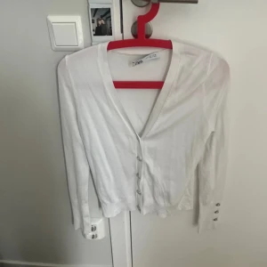 Vit kofta med glittriga knappar från Zara - Supersnygg vit kofta från Zara i storlek S med v-ringning och långa ärmar. Koftan har glittriga knappar framtill och på ärmsluten som ger en extra touch. Perfekt att slänga över en topp för en clean och fräsch look.