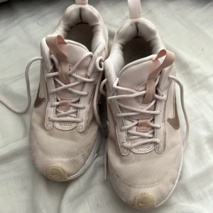 Nike Air Max sneakers i beige och rosa - Snygga Nike Air Max sneakers i ljusbeige med detaljer i rosa och guld. Skorna har mesh- och syntetmaterial, klassisk Air Max-sula och snörning. Perfekta för dig som gillar sportig och trendig stil. Ikonisk Swoosh-logga på sidorna och bekväm passform.