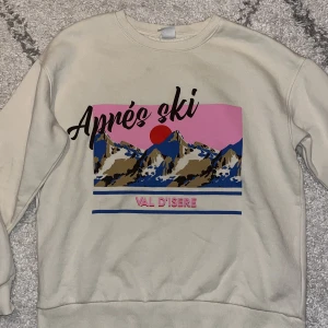Beige sweatshirt med Après ski-tryck - Cool beige sweatshirt från Lindex med stort färgglatt tryck på bröstet som visar berg, sol och texten 'Après ski Val D'Isere' i svart och rosa. Tröjan har rund hals och långa ärmar, perfekt för dig som gillar retrovibbar och grafiska prints.