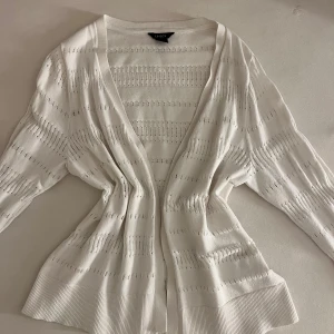Vit stickad kofta från Lindex - Superfin vit stickad kofta från Lindex med lång ärm och öppen front. Koftan har ett diskret hålmönster och breda ribbade muddar vid ärmslut och nederkant. Perfekt att slänga över en topp för en chill och fräsch look.