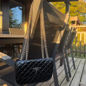 Svart quiltad axelväska från Guess - Säljer en svart quiltad axelväska från Guess med kedjerem i guld och klassisk trekantslogga framtill. Väskan är i glansigt skinnimitation och har ett ljust innerfoder med diskret mönster