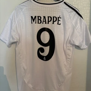 Real Madrid fotbollströja Mbappe 9 - Snygg Real Madrid-tröja med Mbappé på ryggen i storlek Small. Tröjan är i toppskick och knappt använd – perfekt för dig som vill ha en fräsch och stilren supportertröja, vare sig det är till match, träning eller vardagsbruk. Hör gärna av dig vid frågor och passa på att kika in mina andra annonser för fler fynd!