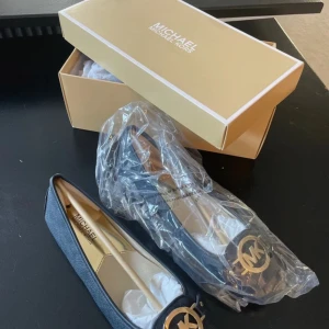 Svarta ballerinaskor Michael Kors - Stilrena svarta ballerinaskor från Michael Kors i skinn med guldfärgad MK-logga framtill. Skorna har rund tå och platt sula, perfekt för en snygg och bekväm look. Klassisk design med exklusiv känsla och diskreta detaljer. Helt nya. Storlek 37-38