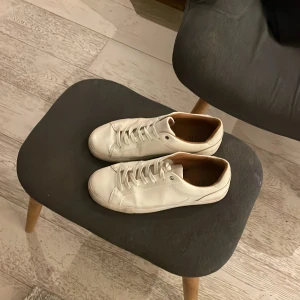 Vita minimalistiska sneakers i skinn - Snygga vita sneakers i stilren design med rund tå och platt sula. Skorna har vita snören och är tillverkade i slätt skinn med ljusbrun insida. Perfekta för dig som gillar en clean och klassisk look.