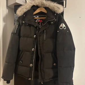 Svart dunjacka med päls från Moose Knuckles - Snygg svart dunjacka från Moose Knuckles med fluffig päls på huvan och coola metalldetaljer. Jackan är i storlek M men sitter mer som en S. Den är rätt använd och säljs pga den inte passar min stil längre.