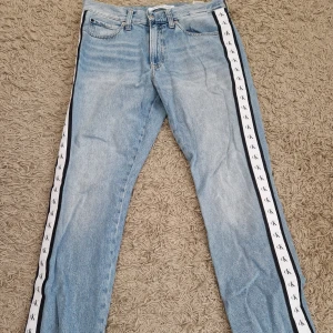 Calvin Klein ljusblå jeans med logoband - Ljusblå jeans från Calvin Klein med raka ben och vita logoband med svarta CK-detaljer längs sidorna. Storlek W31 L32. Byxorna har använts ett fåtal gånger så är i mycket bra skick. Nypris 1000 kr. 