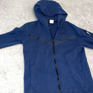 Marinblå Nike hoodie med dragkedja - Snygg marinblå hoodie från Nike med hel dragkedja och huva. Tröjan har svarta detaljer, diskret Nike-logga på bröstet och en praktisk ficka med dragkedja på ärmen. Perfekt för chill eller träning, riktigt skön och stilren.