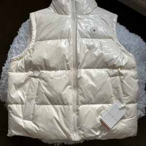 Vit dunväst från Tommy Hilfiger - Säljer en fluffig vit dunväst från Tommy Hilfiger. Vändbar design  med glansig dunyta på ena sidan och mjuk fleece på andra. Hög krage med broderad logga, två fickor och dragkedja framtill. Aldrig använd. prislapp kvar. Sitter mer som en L. Nypris 2600