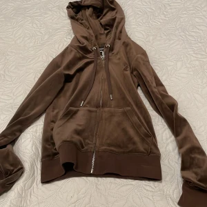 Brun velour hoodie Juicy Couture XS - Säljer en brun hoodie från Juicy Couture i mjuk velour med dragkedja framtill, snörning vid huvan och fickor. Klassisk JC-logga broderad på bröstet. Perfekt för chill dagar och har en skön loose passform med långa ärmar.