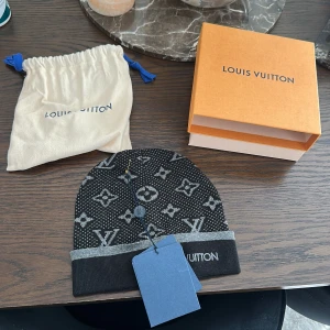 Louis Vuitton svart mössa med monogram - Exklusiv svart mössa från Louis Vuitton med ikoniskt monogrammönster i grått. Mössan har en bredare grå kant nertill och LV-logga broderad på sidan. Kommer med originalförpackning och dustbag. Perfekt accessoar för att lyfta din stil. Helt ny och oanvänd.
