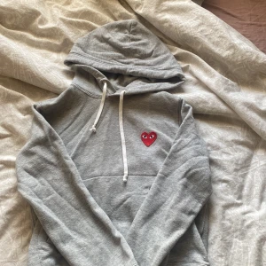 Grå hoodie Comme des Garçons Play - Grå hoodie från Comme des Garçons Play med klassisk röd hjärtlogga med ögon broderad på bröstet. Tröjan har huva med snörning och ficka framtill. Mjuk bomullskvalitet och långärmad modell. Liten i storleken.