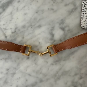 Brunt skärp med guldfärgat spänne från ARKET  - Snyggt brunt bälte i skinn med ett unikt guldfärgat metallspänne i mitten som påminner om ett bett. Bältet har stilrena sömmar längs kanterna och passar perfekt för dig som gillar klassiska detaljer med en twist.