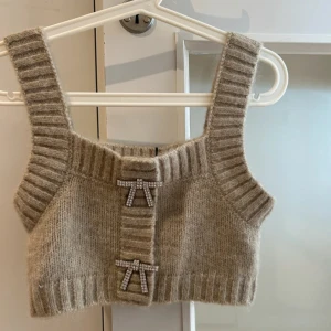 Ny Beige stickad linnetopp med rosetter - Superfin beige stickad linnetopp med breda ribbade axelband och två glittriga rosetter framtill. Mjuk och mysig känsla, perfekt att styla lager på lager eller som den är. Snyggt ribbade detaljer och croppad passform för en trendig look.  Oanvänd 