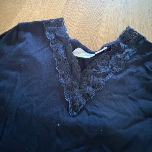 Mörkblå långärmad topp med spets - Mörkblå långärmad topp med snygg v-ringning dekorerad med spetsdetaljer. Toppens material känns mjukt och stretchigt, perfekt för en chill och stilren look. Passar dig som gillar enkla plagg med en twist.