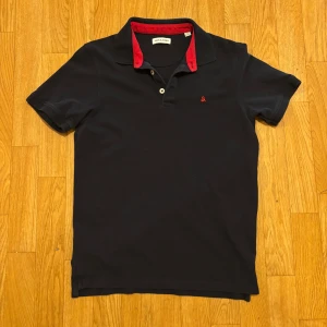 Mörkblå pikétröja från Jack & Jones - Snygg mörkblå pikétröja från Jack & Jones med röd krage och liten röd broderad logga på bröstet. Klassisk piké med korta ärmar, knappar vid halsen och mjukt bomullsmaterial. Perfekt för en clean och stilren look.