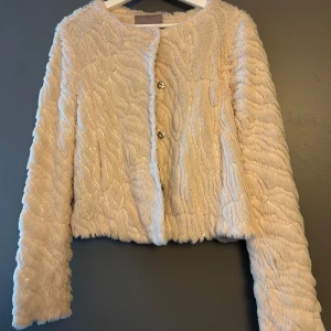 Beige pälsjacka med paljetter Vero Moda - Supermjuk och fluffig beige pälsjacka från Vero Moda med glittriga paljettdetaljer. Jackan har lång ärm, rund halsringning och stängs med två knappar framtill. Perfekt för dig som vill sticka ut med en cozy men ändå glam look.