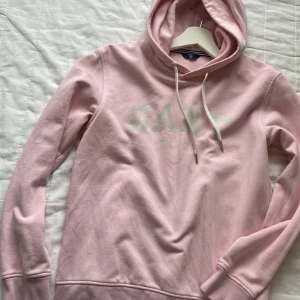 Ljusrosa hoodie från GANT - Superfin ljusrosa hoodie från GANT med vit logga framtill. Tröjan har huva med snörning. Perfekt för en chill och avslappnad stil. Klassisk passform och ribbade muddar vid ärmslut och nederkant. Muddarna har lite tecken på användning, (lite färg som man kan se på bilden) Hoodien är i barn storlek men passar även XS/S
