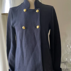 Mörkblå skepparkavaj jacka  med guldknappar - Snygg mörkblå skepparkavaj jacka med ståkrage och dekorativa guldknappar framtill och vid ärmslut. Jackan har en klassisk dubbelknäppt design, raka långa ärmar och diskreta fickor. Materialet är mjukt och känns som ull, perfekt för dig som gillar stilrena och tidlösa plagg.