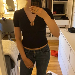 Svart croppad topp från BikBok XS - Svart croppad topp från BikBok med v-ringning och snygg cut-out detalj framtill, dekorerad med en silver ring framtill. Toppen har korta ärmar och är figurnära i modellen. Perfekt för dig som gillar stilrena plagg med en twist.