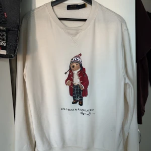 Vit Polo Bear sweatshirt från Ralph Lauren - Vit sweatshirt från Ralph Lauren med ikoniskt Polo Bear-broderi framtill. Tröjan har rund hals, långa ärmar och en relaxed passform. Perfekt för dig som gillar klassisk stil med en twist. Broderiet har björn i röd jacka och mönstrad mössa.