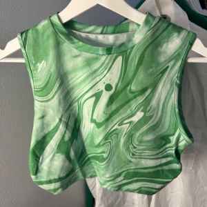 Grön mönstrad croppad linnetopp - Säljer en croppad linnetopp med grönt och vitt swirl-mönster. Toppen är ärmlös och har en tight passform med lätt rynkade detaljer i sidorna. Perfekt för dig som vill sticka ut med en cool och färgstark look. (Endast använd 1 gång) passar både M och S.