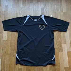 Vintage Nike Tiempo Grå T-Shirt - Vintage Nike Tiempo T-Shirt i funktionsmaterial och färgen grå. OBS! Finns en del sömmar som har lösa trådar, inget har gått upp än men kan eventuellt göra det! Storlek: L, Armhåla till Armhåla: 55 cm, Längd: 74 cm, Halsöppning till Armöppning: 44 cm.  Material: 100% Polyester