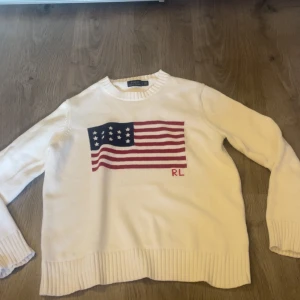 Ralph Lauren tröja  - Vit stickad tröja från Polo Ralph Lauren med amerikansk flagga i rött, blått och vitt på bröstet samt RL-broderi. Rund hals och ribbade muddar vid ärmslut och nederkant. 