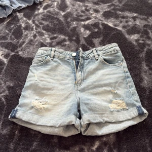 Ljusblå jeansshorts med slitningar H&M - Ljusblå jeansshorts från H&M med avslappnad passform och uppvikta benslut. Shortsen har slitna detaljer framtill, klassiska fem fickor och stängs med dragkedja och knapp. Perfekta för sommardagar och enkel att matcha med allt.