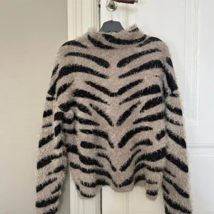 Zebramönstrad fluffig tröja NA-KD - Supermjuk och fluffig tröja från NA-KD i beige och svart zebramönster. Tröjan har hög hals och långa ärmar, perfekt för att sticka ut med en cool look. Är oversized 🥰