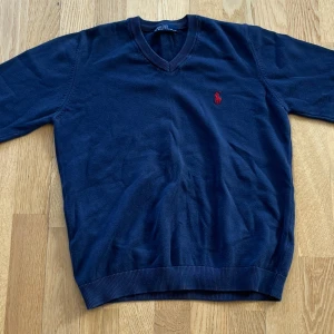 Marinblå stickad tröja Polo Ralph Lauren - Snygg marinblå stickad tröja från Polo Ralph Lauren med klassisk v-ringning och röd broderad logga på bröstet. Tröjan har ribbade muddar och är perfekt för dig som gillar stilren och tidlös design.