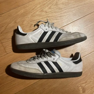 Adidas samba adv - Klassiska Adidas Samba sneakers i vitt skinn med svarta detaljer och de ikoniska tre ränderna på sidorna. Låg modell med snörning och mörk gummisula. Perfekta för dig som gillar retrostil och streetwear.