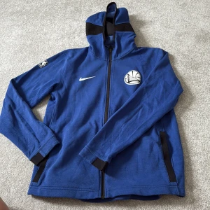 Blå Nike NBA zip hoodie Warriors - Blå zip hoodie från Nike med Golden State Warriors-logga på bröstet och NBA-märke på ärmen. Jackan har hög krage, svarta detaljer och två fickor. Tillverkad i mjukt polyester Dri-Fit material som andas. Perfekt för basketfans som vill ha sportig stil.