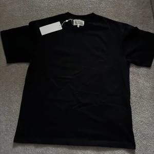 Maison Margiela svart t-shirt  - Svart t-shirt från Maison Margiela med ikoniskt vitt tryck och siffror på ryggen. Klassisk rund hals och korta ärmar. Tillverkad i mjuk bomull, perfekt för dig som gillar stilrena designer med edge. Made in Italy och har Margielas signaturtag.