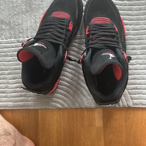 Nike Air Jordan 4 Svart/Röd - Säljer ett par Nike Air Jordan 4 i svart och rött med klassisk Jumpman-logga på hälen. Skorna har meshpaneler, synlig Air-sula och coola detaljer i rött. Ovandelen är i mocka och syntet, med svarta snören och en chunky siluett som sticker ut. Dem har använts ett par gånger 