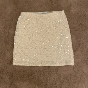 Paljettkjol i vitt från Lindex - Snygg vit kortkjol från Lindex med glittriga paljetter över hela ytan. Kjolen har en rak passform och är perfekt för dig som vill sticka ut med lite extra bling. Mjuk insida och elastisk midja för skön känsla. Passar dig som gillar att glänsa!