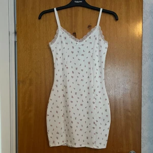 Vit blommig klänning - Supersöt vit figurnära klänning med små rosa blommor från H&M Divided i strl S. Klänningen har smala reglerbara axelband, spetsdetaljer vid v-ringningen och en liten rosett framtill. Aldrig använd!💞