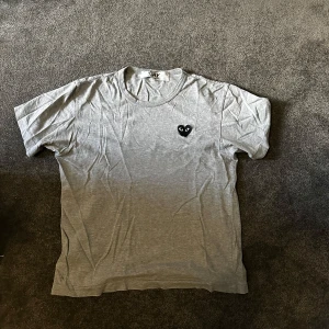 Grå t-shirt Comme des Garçons Play - Grå t-shirt från Comme des Garçons Play i mjuk bomull. Klassisk rund hals och korta ärmar. På bröstet sitter det ikoniska svarta hjärta med ögon. Enkel och clean design som funkar till allt. Perfekt för dig som gillar stilrena plagg med en twist.