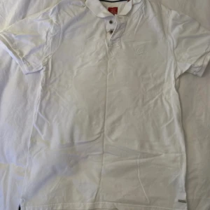 Vit pikétröja  - Snygg vit pikétröja med broderad logga på bröstet och metallmärke vid nederkanten. Klassisk krage och två knappar framtill. Perfekt för dig som gillar stilrena och enkla plagg. Materialet är mjuk bomull och modellen har korta ärmar.