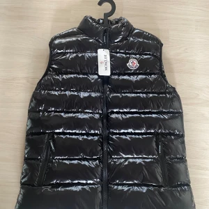 Svart dunväst från Moncler - Snygg svart dunväst från Moncler med glansig finish och klassisk logga på bröstet. Västen har hög krage, två dragkedjefickor och tydlig Moncler-detalj på dragkedjan. Perfekt för lager-på-lager och streetstyle.