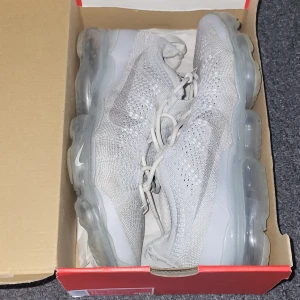 Nike Vapormax Flyknit vita sneakers - Nike Vapormax Flyknit sneakers i helvitt med stickad ovandel och genomskinlig, bubblig sula. Skorna har snörning och en sportig vibe, perfekt för dig som gillar streetstyle. Lätta och luftiga med modern design och diskret Swoosh på sidan.