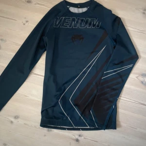 Marinblå  långärmad träningströja Venum - Säljer en svart långärmad träningströja från Venum med tryckt logga och grafiska linjer i grått och svart. Tröjan är i syntetmaterial och har en tight passform, perfekt för gym eller kampsport.