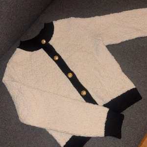 Beige kofta med svarta detaljer från H&M - Säljer en beige/svart kofta från H&M med svarta ribbade kanter och  guldknappar. Koftan har långärmad passform och är stickad. Perfekt för dig som gillar klassisk stil. Använd 3-4 gånger. Nytt skick! 